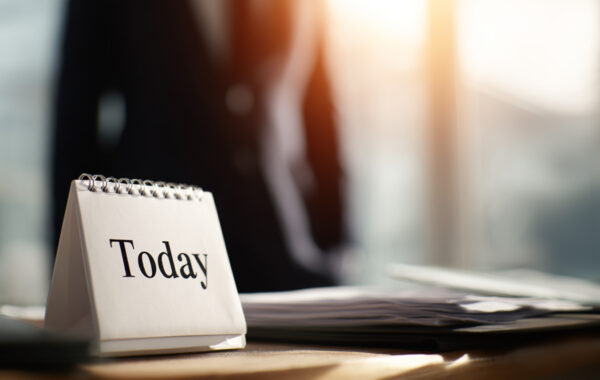 Das Bild zeigt einen Abrisskalender. Im Hintergrund verschwommen ein Geschäftsmann im Büroraum.